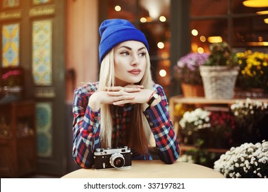 Portrait of  beautiful stylish young woman sitting in street cafe and drinking coffee. Hipster with old retro camera. Model looking aside. Lights and flowers background. City lifestyle. Toned

