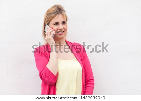 Portrait of beautiful successful young woman in pink blouse standing, using and making call on her smartphone with happy face and talking with friend. Indoor, isolated, studio shot, white background