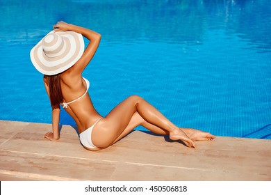 Portrait of beautiful tanned sporty slim woman relaxing in swimming pool spa. Creative white hat and bikini. Hot summer day and bright sunny light.