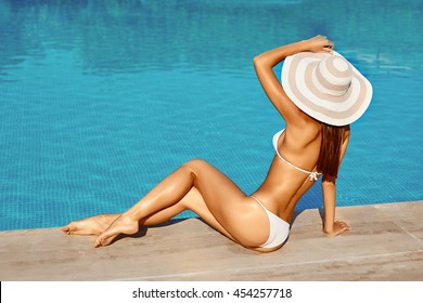 Portrait of beautiful tanned sporty slim woman relaxing in swimming pool spa. Creative white hat and bikini. Hot summer day and bright sunny light.