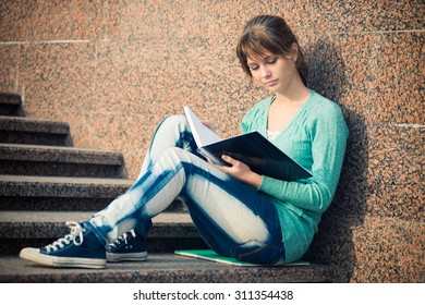 Portrait Of A Beautiful Teenage Student Girl Sitting On Stairs In Park With Copy Book And Studing. Sunny Summer Day. Modern And Casual Lifestyle. Woman With Note Pad.