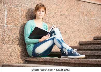 Portrait Of A Beautiful Teenage Student Girl Sitting On Stairs In Park With Copy Book And Studing. Sunny Summer Day. Modern And Casual Lifestyle. Woman With Note Pad.