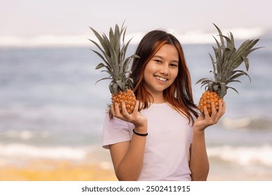 Portrait of beautiful teenager girl holding two whole pineapples. Smiling face. Blurred sandy beach, ocean and sky background. Organic fruit concept. Healthy lifestyle. Pandawa beach, Bali, Indonesia - Powered by Shutterstock