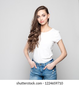 Portrait of beautiful teenager girl wearing white t-shirt and jeans, smiling at camera. Standing with hands in pockets against grey background. 