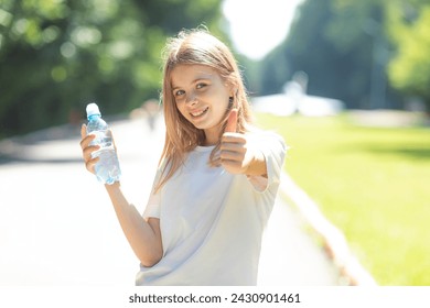 Portrait of beautiful teenager pretty girl, young thirsty woman is drinking pure fresh water from bottle at warm sunny summer day outdoors, holding bottle in hand and showing thumbs up - Powered by Shutterstock