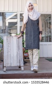 Portrait Of Beautiful Thai Young Muslim Woman Coffee Shop Owner Wearing Hijab Standing At The Entrance Of His Coffee Shop With A Wooden Board. Barista Standing In The Doorway Of A Restaurant.