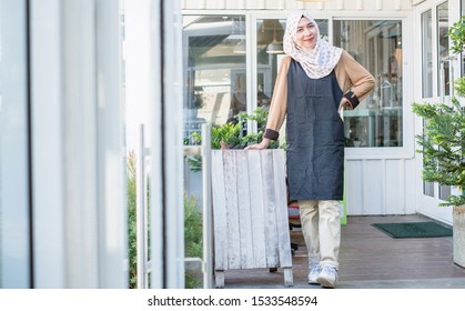 Portrait Of Beautiful Thai Young Muslim Woman Coffee Shop Owner Wearing Hijab Standing At The Entrance Of His Coffee Shop With A Wooden Board. Barista Standing In The Doorway Of A Restaurant.