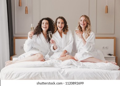 Portrait Of Beautiful Three Women 20s Wearing Bathrobe Lying In Big Bed In Posh Apartment Or Hotel Room With Glasses Of Champagne During Bachelorette Party