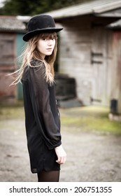 Portrait Of A Beautiful Victorian Style Woman Wearing A Bowler Hat And Black Dress Standing In Front Of Country Farm Stables