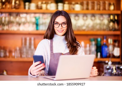 Portrait of a beautiful waitress text messaging and using laptop while standing behind the counter at a restaurant. Small business. 