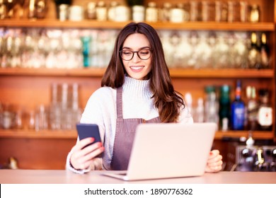 Portrait of a beautiful waitress text messaging and using laptop while standing behind the counter at a cafe. Small business. 