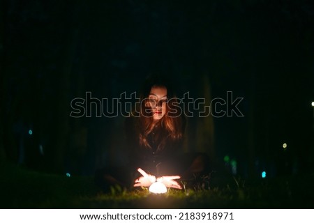 Portrait of beautiful witch in black dress with magic fire ball in forest at dark night