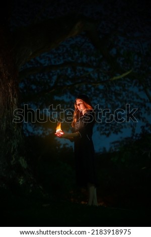 Portrait of beautiful witch in black dress with magic fire ball in forest at dark night