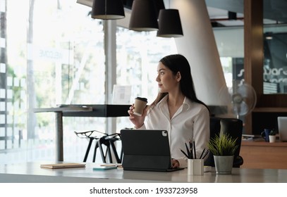 Portrait Of Beautiful Woman Or Accountant Sitting At Desk In Modern Office With Interior Drinking Hot Beverage Holding Cup With Coffee In Hands Looking Outside Enjoying Freetime.