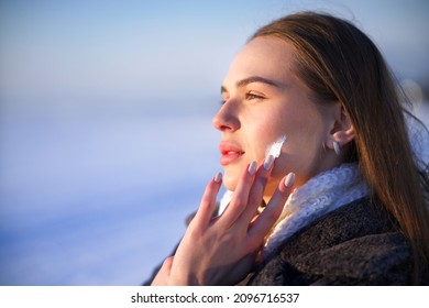 Portrait Of Beautiful Woman Applying Cosmetic Skin Cream On Face And Hands, Protecting From Cold Weather At Winter Season. Pretty Girl Take Care Of Her Skin Outdoors On Snowy Nature Background. 