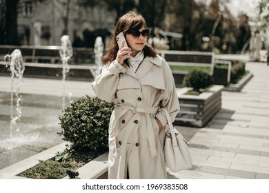 Portrait Of Beautiful Woman In Beige Trench Coat, Sunglasses Walking In The City Near The Fountains And Talking On The Phone. Stylish Adult Mother. Business Woman. Blogger, Consultation, Busy, Old