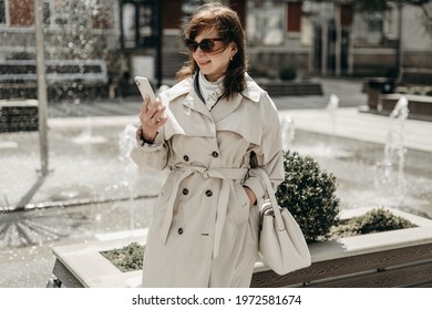 Portrait Of Beautiful Woman In Beige Trench Coat, Sunglasses Walking In The City Near The Fountains And Talking On The Phone. Stylish Adult Mother. Business Woman. Blogger, Consultation, Busy, Old