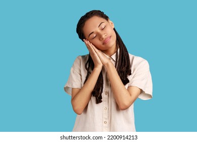 Portrait Of Beautiful Woman With Black Dreadlocks Sleeping Laying Down On Her Palms, Having Comfortable Nap And Resting, Dozing Off, Wearing White Shirt. Indoor Studio Shot Isolated On Blue Background