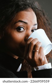 Portrait of Beautiful Woman with black skin and long wavy hair, elegantly dressed, drinking coffee from a white cup. She has an amused and intelligent expression, almost intrigued, but sympathetic.  - Shutterstock ID 2266959559