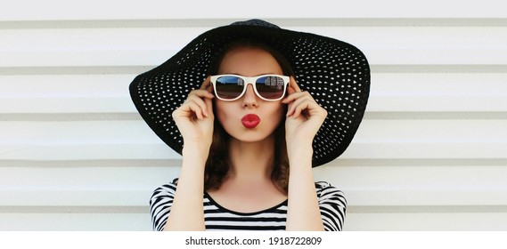 Portrait of beautiful woman blowing her red lips sending sweet air kiss wearing a black round summer straw hat on a white background