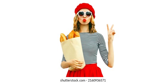 Portrait Of Beautiful Woman Blowing Her Red Lips Sending Sweet Air Kiss Holding Grocery Shopping Paper Bag With Long White Bread Baguette Wearing French Red Beret Isolated On White Background