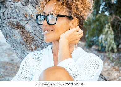 Portrait of a beautiful woman with curly hair gathered, wearing stylish sunglasses and smiling while looking away. She enjoys a serene and happy moment, relaxing alone in a park and embracing a joy - Powered by Shutterstock