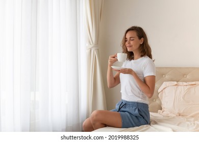 Portrait of beautiful woman drinking morning coffee on bed by the window. Happy beautiful pretty woman relaxing drinking and looking at cup of hot coffee or tea in bedroom. Woman drinking hot coffee.