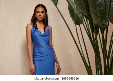 Portrait Of Beautiful Woman Fashion Model Brunette Hair Wear Blue Silk Dress Accessory Jewelry Earrings Cosmetic Face Clothes For Romantic Date Party Walk Summer Collection Tanned Skin Green Leaves.