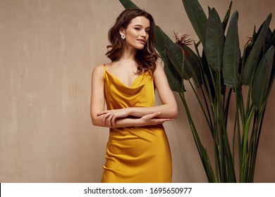 Portrait Of Beautiful Woman Fashion Model Brunette Hair Wear Yellow Silk Dress Accessory Jewelry Earrings Cosmetic Face Clothes Romantic Date Party Walk Summer Collection Tanned Skin Green Leaves.