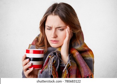 Portrait Of Beautiful Woman Has Headache, Has Bad Cold, Drinks Hot Tea Or Coffee, Wrapped In Checkered Blanket, Looks Miserable, Isolated Over White Concrete Background. Disease And Illness Concept