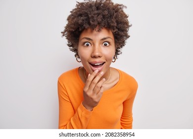 Portrait of beautiful woman hears interesting rumor gazes amazed looks intrigued keeps mouth widely opened dressed in casual orange jumper isolated over white background hears details of smth