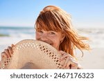 Portrait of beautiful woman hiding behind a straw hat at beach and looking at camera with beautiful green eyes. Closeup face of smiling girl with freckles and red hair relaxing at seaside.