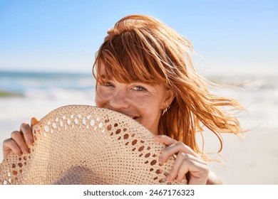 Portrait of beautiful woman hiding behind a straw hat at beach and looking at camera with beautiful green eyes. Closeup face of smiling girl with freckles and red hair relaxing at seaside.
