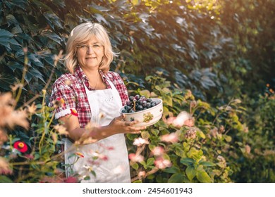 Portrait of a beautiful woman holding black grapes in a bowl outdoors. Senior female standing in the garden with a fruit in her hands. - Powered by Shutterstock