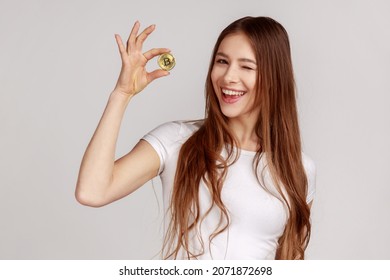 Portrait of beautiful woman holding golden bitcoin, showing btc coin and winking at camera with positive expression, wearing white T-shirt. Indoor studio shot isolated on gray background.