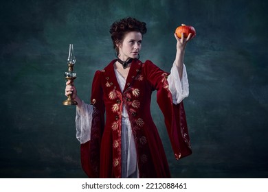 Portrait Of Beautiful Woman In Image Of Medieval Vampire Holding Candle And Pumpkin Isolated Over Dark Green Background. Horror. Concept Of Halloween, Mystery, Gothic Art, Fantasy, Surreal Characters