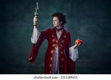 Portrait Of Beautiful Woman In Image Of Vampire Holding Candle And Pumpkin Isolated Over Dark Green Background. Spooky Season. Concept Of Halloween, Mystery, Gothic Art, Fantasy, Surreal Characters