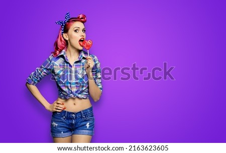 Portrait of beautiful woman licking heart shape lollipop dressed in pinup plaid shirt, isolated over violet background. Purple red girl in retro fashion and vintage studio concept. Tasty sales prices!