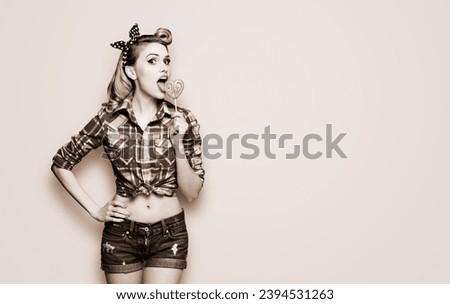 Portrait of beautiful woman licking heart shape lollipop dressed in pinup plaid shirt. Pin up girl in retro fashion and vintage studio concept. Brown toned black and white bw photo