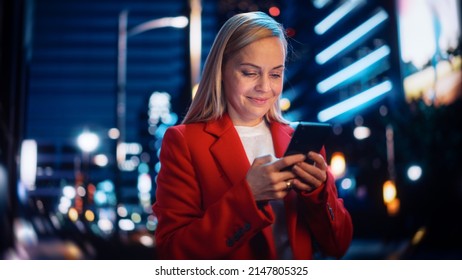 Portrait of a Beautiful Woman in Red Coat Walking in a Modern City Street with Neon Lights at Night. Attractive Female Using Smartphone and Looking Around the Urban Cinematic Environment. - Powered by Shutterstock