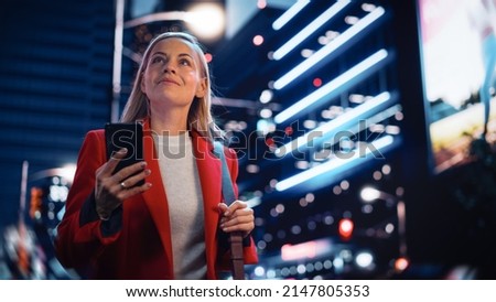 Portrait of a Beautiful Woman in Red Coat Walking in a Modern City Street with Neon Lights at Night. Attractive Female Using Smartphone and Looking Around the Urban Cinematic Environment.