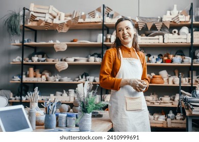 Portrait of a beautiful woman running a pottery studio. Smiling woman potter in apron against background of rack with clay products in workshop - Powered by Shutterstock