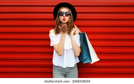 Portrait of beautiful woman with shopping bags blowing her lips sending sweet air kiss wearing black round hat on red background - Shutterstock ID 2068318034