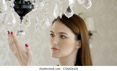 A portrait of a beautiful woman staring at luxurious chandelier and touching the crystals of vintage lamp.