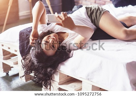 Portrait of beautiful woman waking up in her bed and looks into the phone. Check social networks, send sms. The girl is wearing a T-shirt.