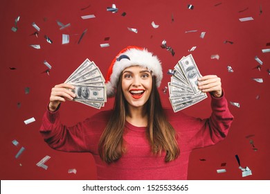 Portrait of beautiful woman wearing Santa Claus red costume smiling and holding fan of money in dollar banknotes isolated over red background.