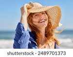 Portrait of beautiful woman wearing straw hat at beach and looking at camera with a big smile. Closeup face of attractive smiling girl with freckles and red hair. Happy young woman enjoing summer.