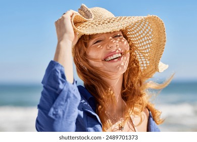 Portrait of beautiful woman wearing straw hat at beach and looking at camera with a big smile. Closeup face of attractive smiling girl with freckles and red hair. Happy young woman enjoing summer.