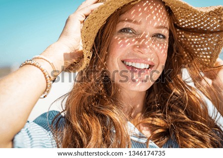 Portrait of beautiful woman wearing straw hat with large brim at beach and looking at camera. Closeup face of attractive smiling girl with freckles and red hair. Happy mature woman enjoying summer.