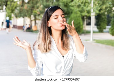 Portrait Of Beautiful Woman In White Shirt And Hair Bezel With Mouth Full Of Food Licking Her Fingers Outdoor In City Park And Enjoying Junk But Tasty Fast Food While Walking